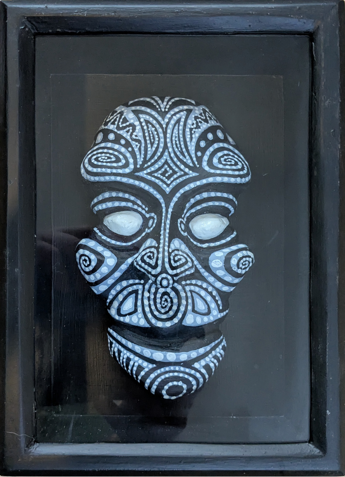 Abner Harris - Black Headie - A three-dimensional, hand-painted plaster sculpture of a stylized head featuring intricate, white-on-black pointillist and scrollwork patterns. The mask-like form has glowing white eyes and highly detailed tribal geometric motifs. The piece is set against a solid black background and professionally housed in a sleek, matching black wooden frame under protective glass, creating a sophisticated graphic noir aesthetic.