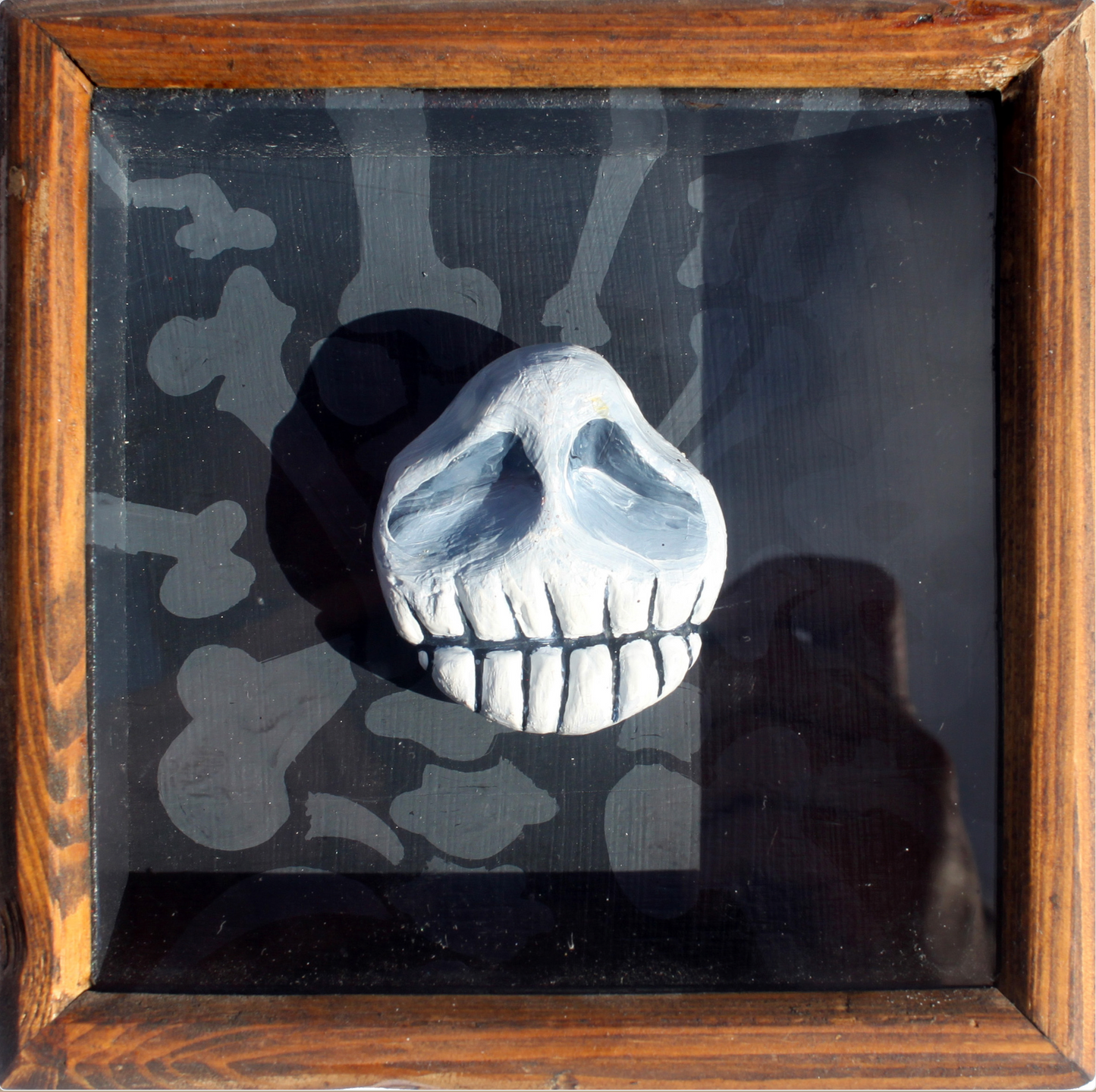 Abner Harris - Bone Headie - A three-dimensional, hand-painted plaster sculpture featuring a stylized, grinning skull with prominent teeth and hollowed eyes. The skull is mounted against a dark background adorned with a ghostly pattern of grey bones. This tactile piece is professionally encased in a rustic, natural grain wooden frame under protective glass, offering a modern, macabre twist on folk art sculpture.