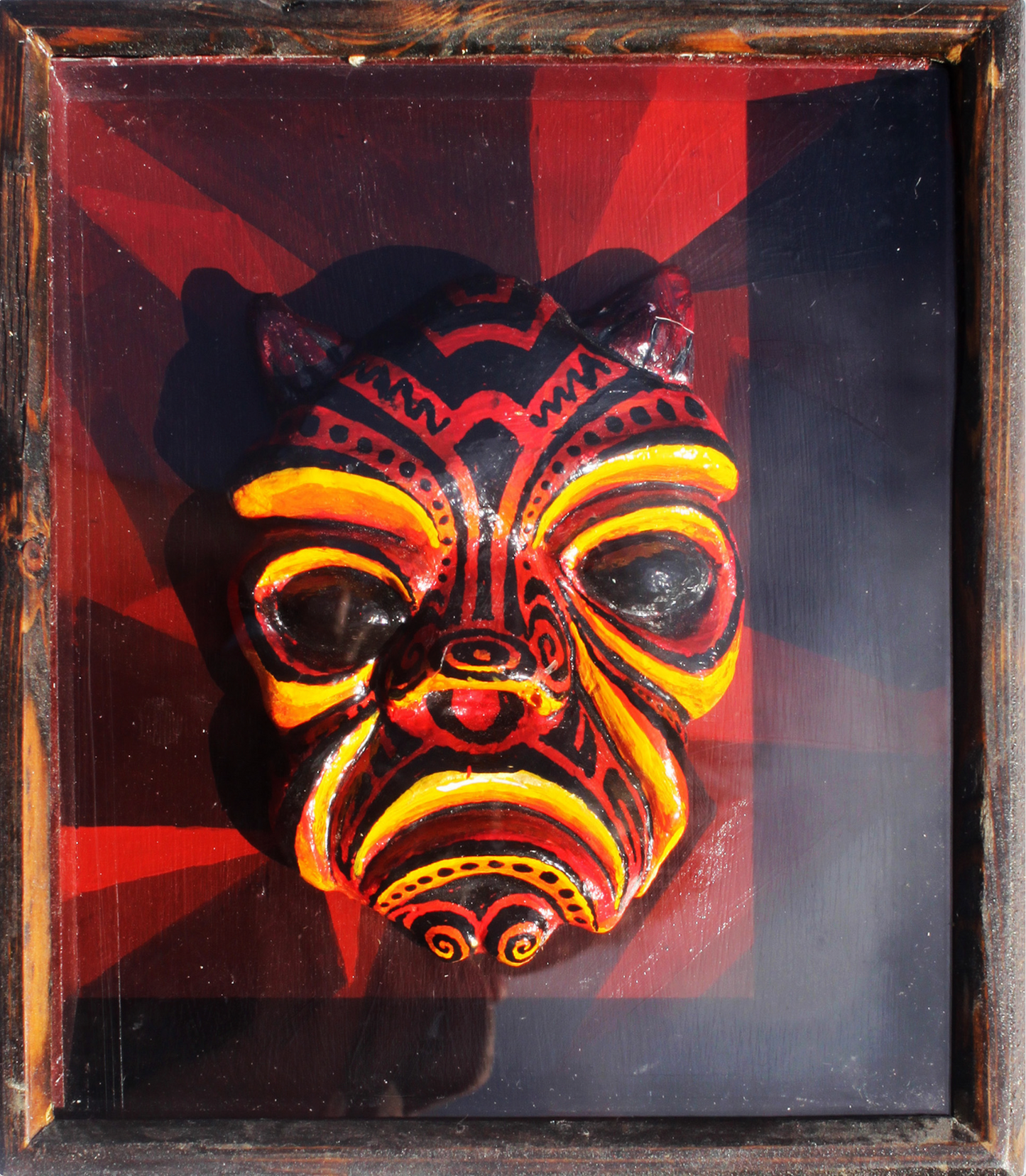 Abner Harris - Fire Headie - A three-dimensional, hand-painted plaster sculpture of a fierce, horned visage. The head features intricate, tribal-inspired patterns in vibrant red, orange, and yellow over a deep black base. It is mounted against a dramatic red and black radiating sunburst background and encased in a dark, char-finished wooden frame under protective glass. This piece combines high-energy color with a bold, sculptural folk-art aesthetic.