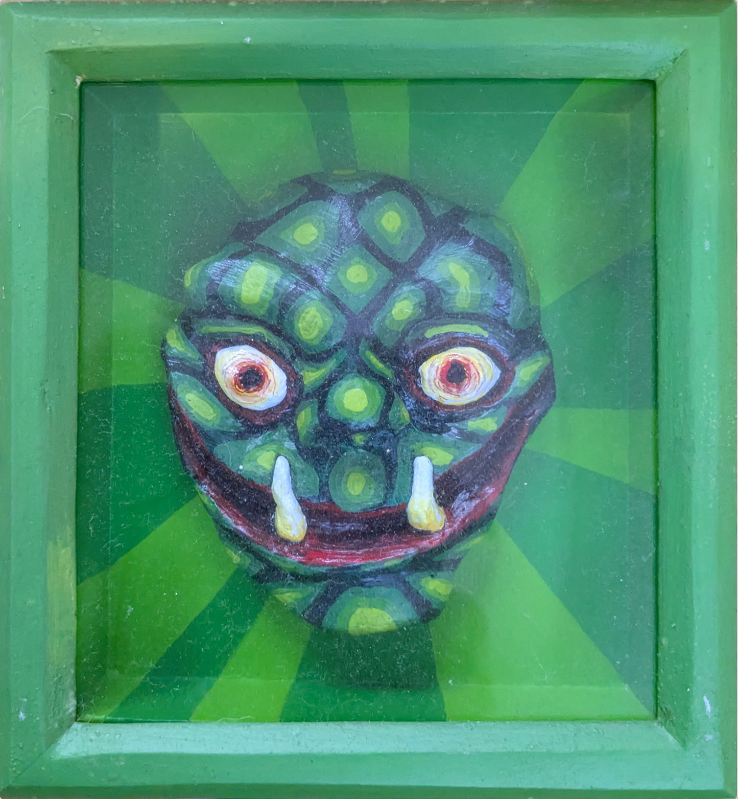Abner Harris - Reptile Headie - A three-dimensional, hand-painted plaster sculpture of a stylized reptilian head with wide, intense eyes and protruding white tusks. The mask-like form features intricate green scales and a patterned crown, set against a radiating two-tone green sunburst background. The piece is professionally housed in a matching lime-green wooden frame under protective glass, blending sculptural folk art with a modern pop aesthetic.